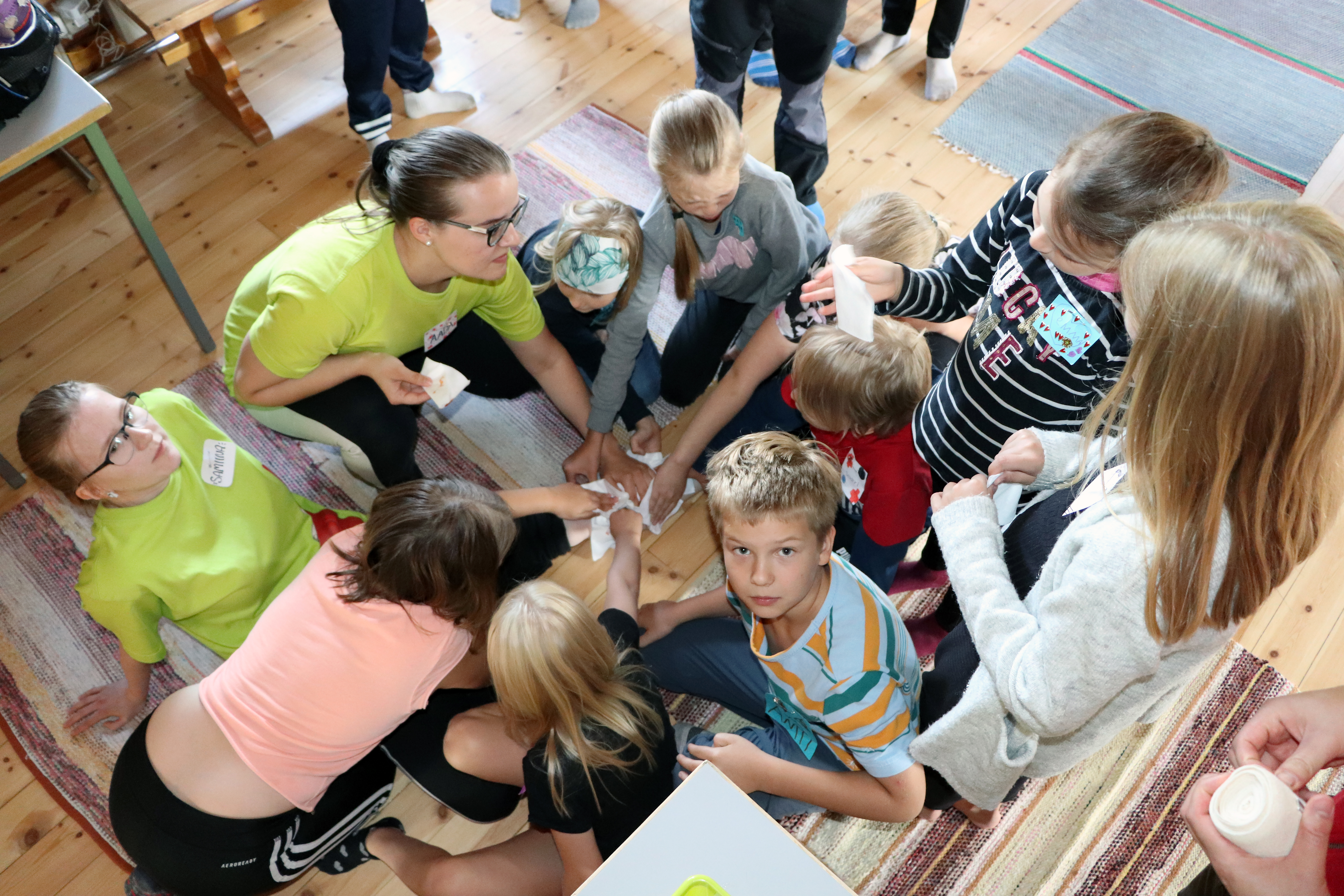 <p>Leiriläiset opettelevat haavan oikeaoppista ensiapua, kun ohjaaja Samiina Rantala sai  ”ison haavan” jalkaansa. Aada Satokangas opastaa lapsia tyrehdyttämään verenvuotoa ja tekemään siteen haavan ympärille. </p>