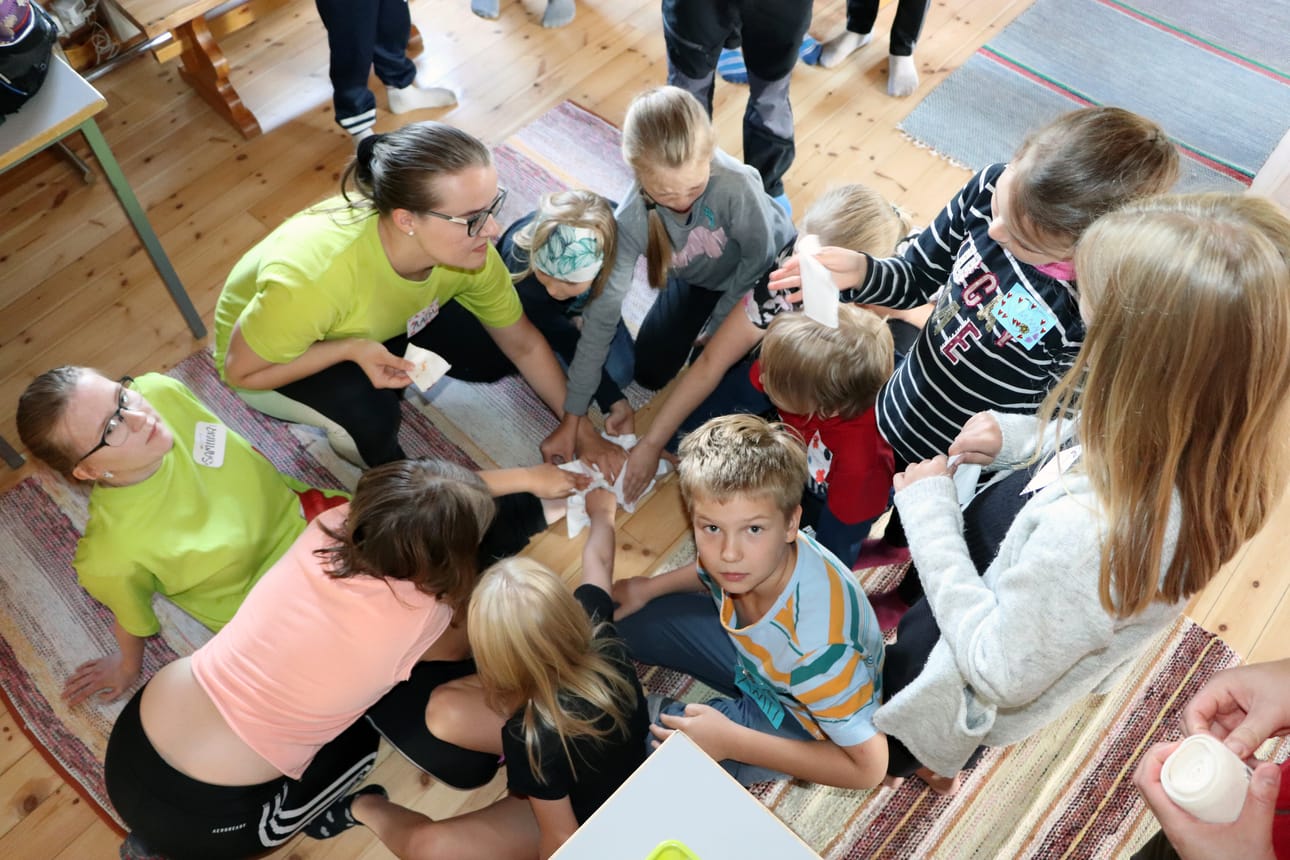 Leiriläiset opettelevat haavan oikeaoppista ensiapua, kun ohjaaja Samiina Rantala sai ”ison haavan” jalkaansa. Aada Satokangas opastaa lapsia tyrehdyttämään verenvuotoa ja tekemään siteen haavan ympärille.