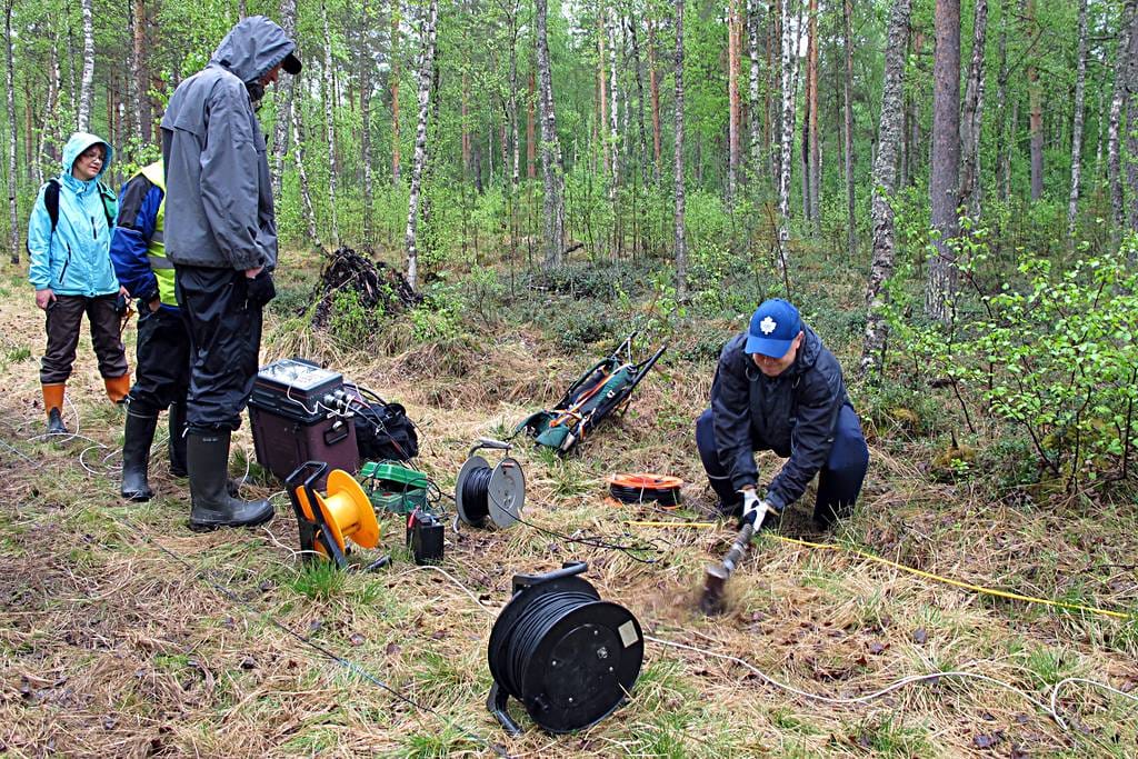 Johtava tutkija Jarkko Okkonen (oik) iskee lekalla metallilevyä, josta lähtevän seismisen aallon avulla tarkastetaan laitteiden toimivuus. Mittareita seuraamassa (vas.) tutkija Helena Vikstedt, geofyysikko Juha Mursu ja erikoistutkija Kari Moisio.