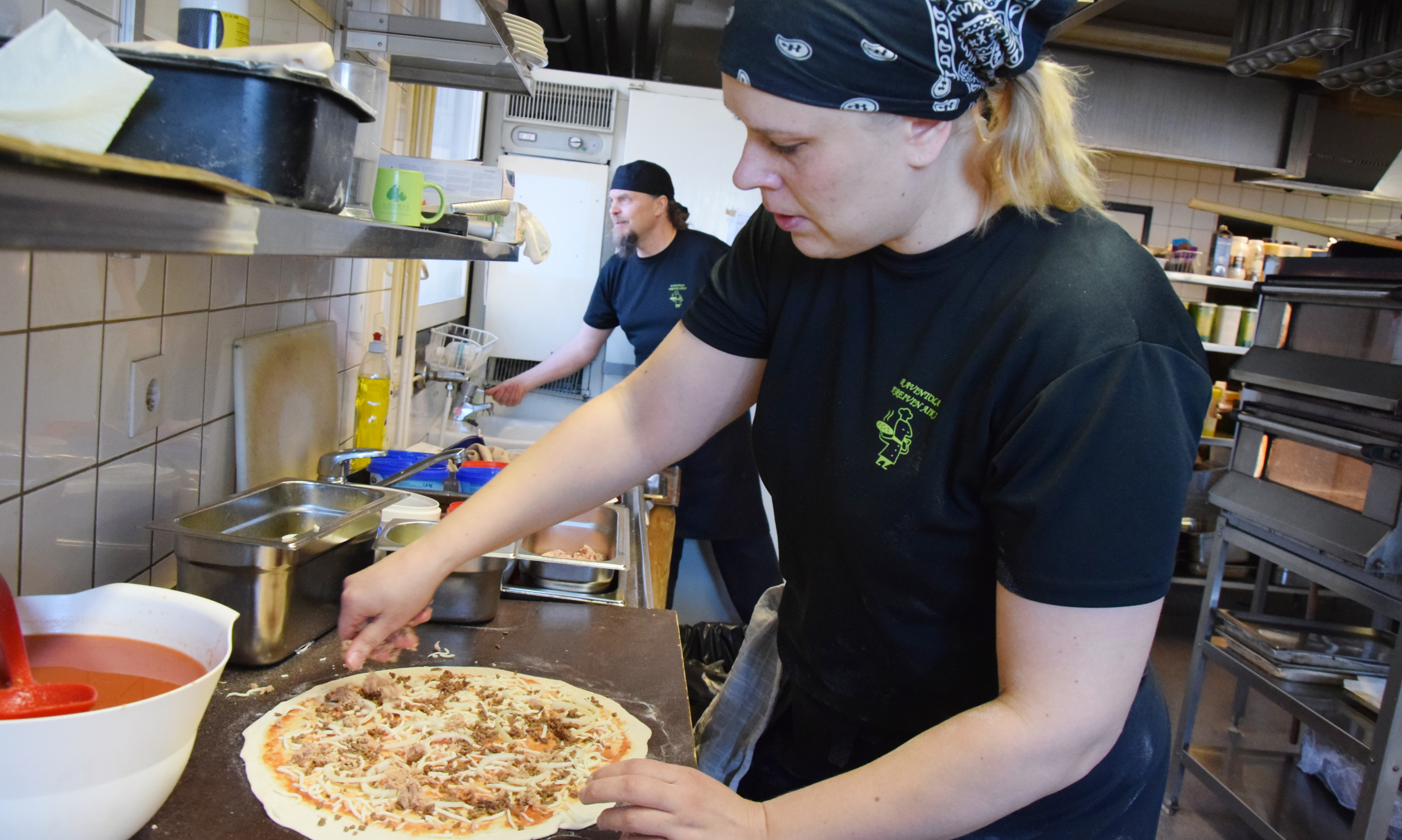 Maria Pääkkö täyttää ohueksi kaulittua pizzaa. Taustalla Kreivinajan yrittäjä Antti Ruonala.