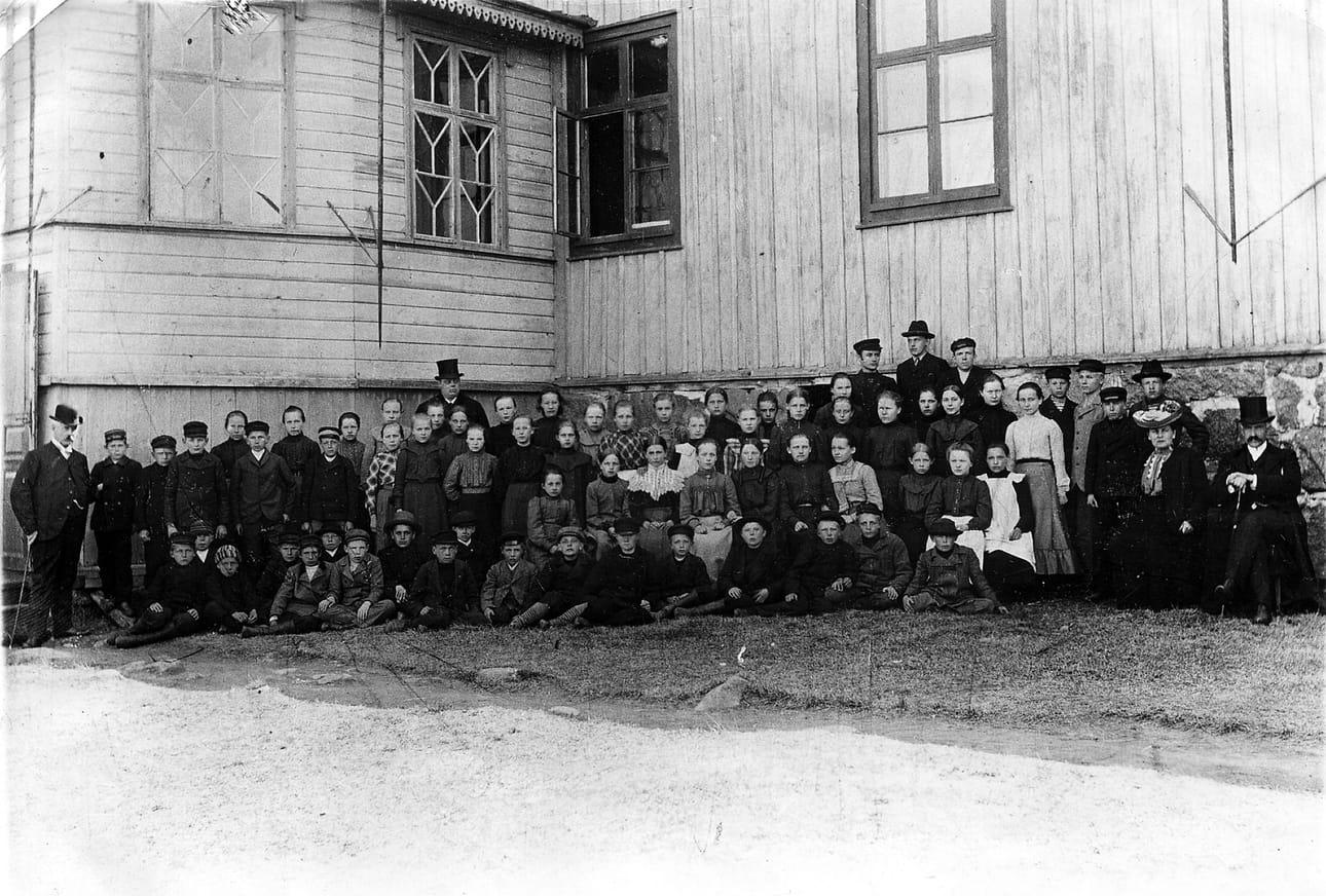 Vanha alkeiskoulu tuhoutui tulipalossa vuonna 1908, kun tulipalo tuhosi kirkon ja sen kupeessa sijainneen koulun. Kuvassa vanhan alkeiskoulun opettajia ja oppilaita koulunsa edustalla. Taustalla silinteripäinen rehtori Hugo Ingman, vasemmassa reunassa knallipäinen opettaja G. A. Wangell ja oikeassa reunassa rouva Beata Ingman ja opettaja Dudley Campbell. Kuva: Raahen museon kokoelmista.