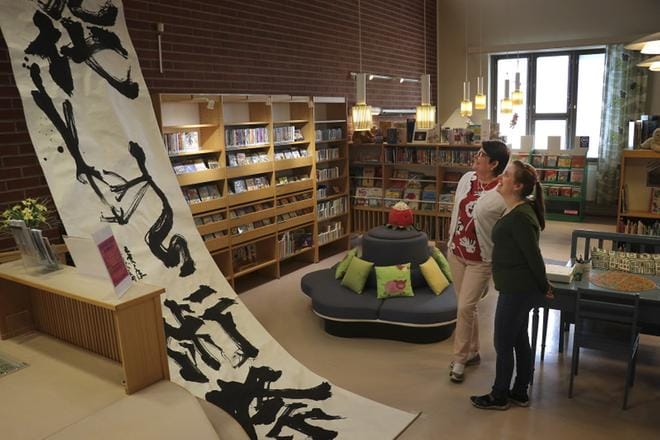 Kirjastotoimenjohtaja Tarja Kyrönlahti ja kirjastosihteeri Kerttu Anttiroiko-Rauma miettivät isolle teokselle sopivaa sommittelua. Kannen kuvassa Elina Alkio esittelee näyttelyä pystyttämässä olleille Kudo Shuheille (vas.), Magori Bumpeille, Fujiyama Toyolle suomalaisia tervehdyksiä. KUVAT: JOHANNA KATAJA