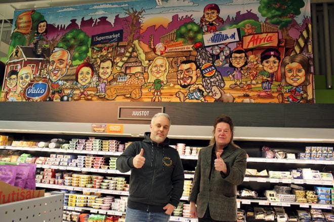 Kauppias Olli Pehkonen (vas.) ja karikatyyritaiteilija Jorma Lampela ovat tyytyväisiä Asterixin gallialaiskylään pohjautuvaan teokseen, joka on nostettu juustohyllyjen yläpuolelle. Monet asiakkaat pysähtyvät tunnistamaan kuvan henkilöitä. KUVA: KIRSTI MÄKELÄ