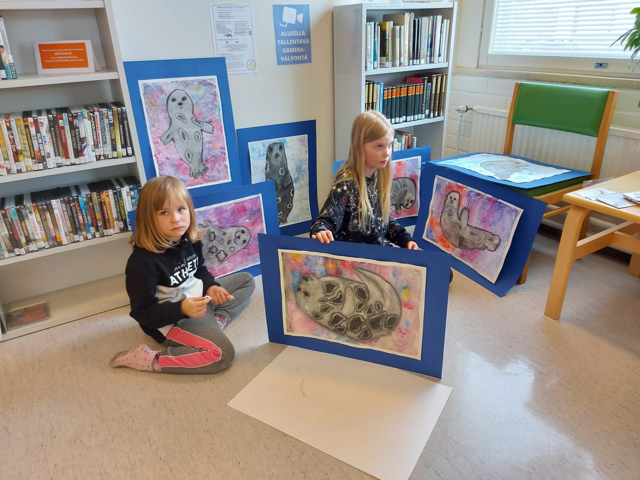 Viivi Haakana ja Iida Ohenoja valmistautuvat sommittelemaan leppoisia norppateoksia kirjaston seinälle. Jokihelmenopiston kuvataidekoulun kevätnäyttely on esillä Piippolan kirjastolla 28.4. saakka.Kahdeksasluokkalainen Jenny-Reeyta Äijälä on käynyt kuvataidekoulua ekaluokasta saakka. Nyt hän sai valmiiksi kuvataiteen perusopetuksen näyttötyönsä "Colors in the Darkness".Emma, Anni ja Kerttu valmistelevat kultaista alustaa näyttelyyn tuoduille töilleen. Ihmeellisiä metsiä esittävät dioraamat sopivat hienosti satusienten taustaksi villien villapaitojen katveessa.