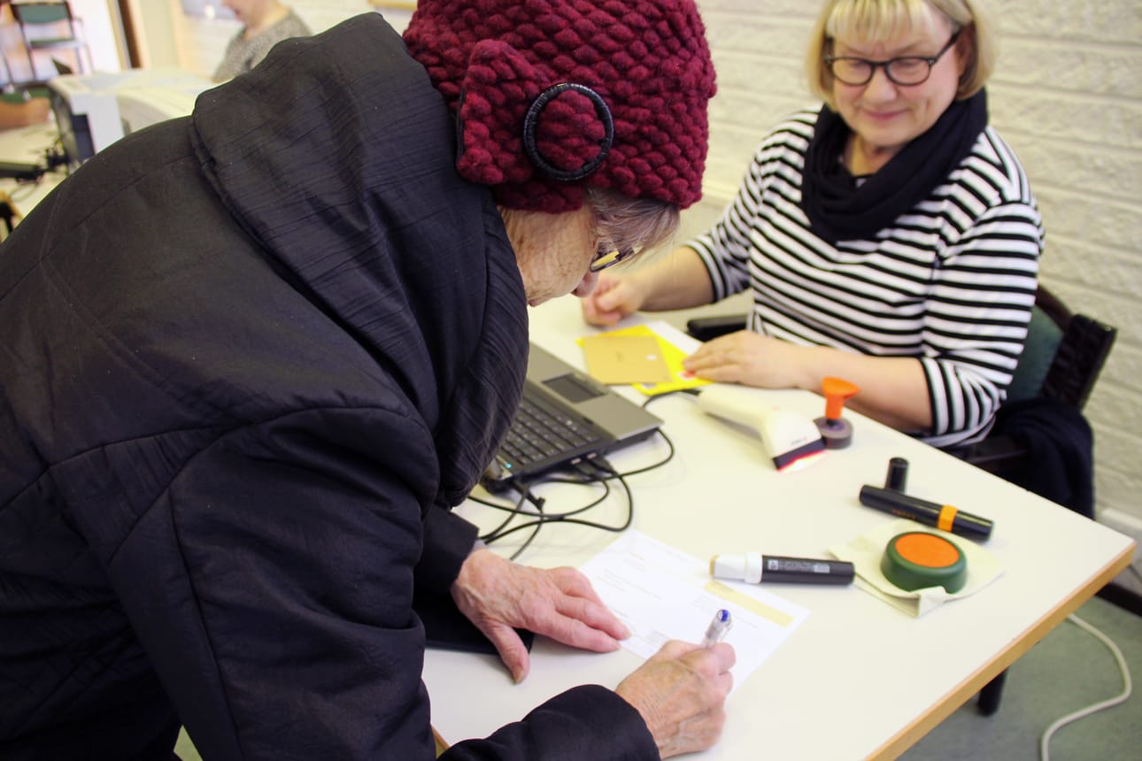 Ritva Luomajoki allekirjoittaa ennakkoäänestyksen lähetekirjettä. Taustalla vaalivirkailija Taina Stoor.