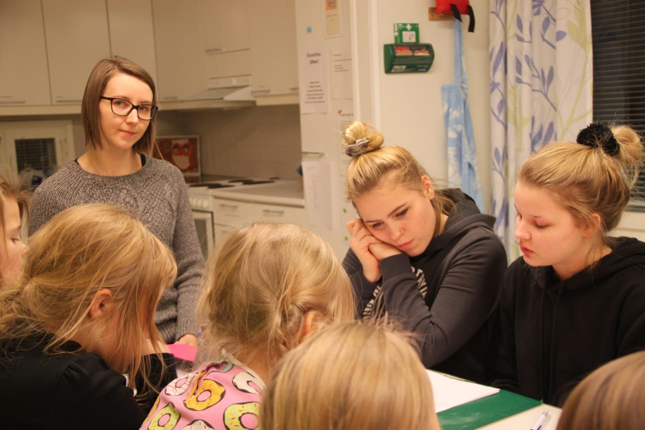 Pöydän päässä istuva Eva-Maria Mustonen ja kokkikerhon ohjaajat Anna Kuukasjärvi ja Anna-Riikka Wikstedt kertovat tytöille, miten lämpimiä leipiä valmistetaan.