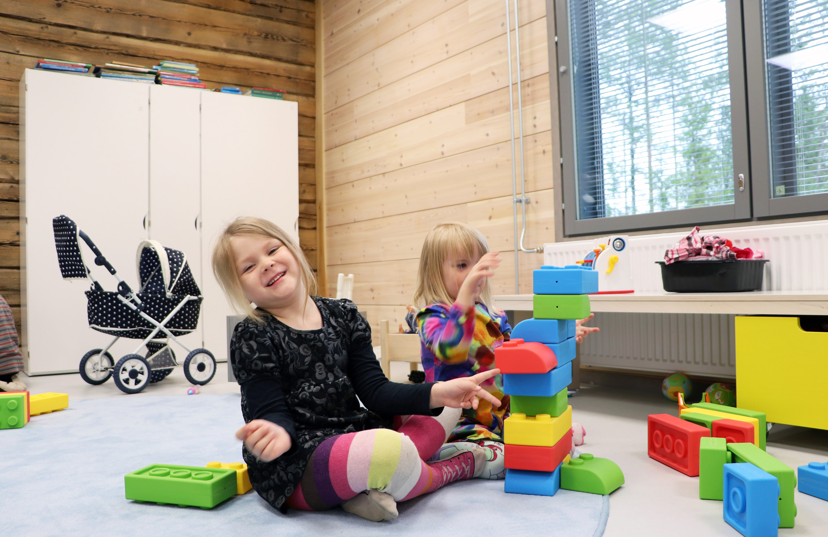 Titta Riepula ja Aune Jussila ovat Kipinän ryhmiksessä päivähoidossa. Laajennusosan avarat ikkunat tuovat valoa leikkihuoneeseen. Taustalla näkyy vanhaa hirsipintaa, joka oli ennen koulurakennuksen päätyseinä.