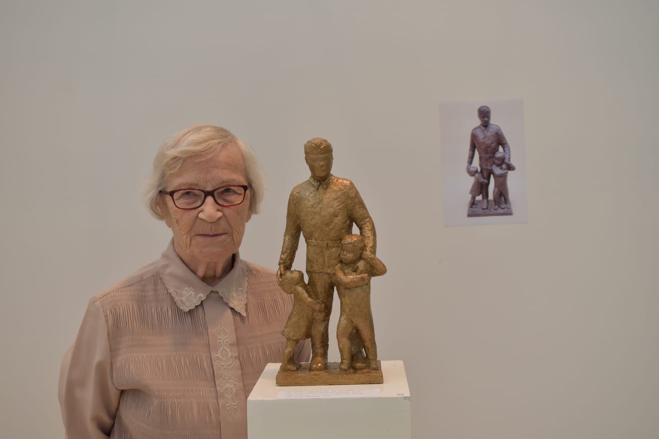 Marjatta Vilhunen teoksensa "Et lähtis isä" vierellä. Vilhusen oma isä kaatui sodassa vuonna 1941. Veistos on lakattua kipsiä, sen takana on kuva veistoksen pronssiversiosta. Teos on yksi Vilhusen näyttelyn vanhimpia, sillä se on vuodelta 1998.