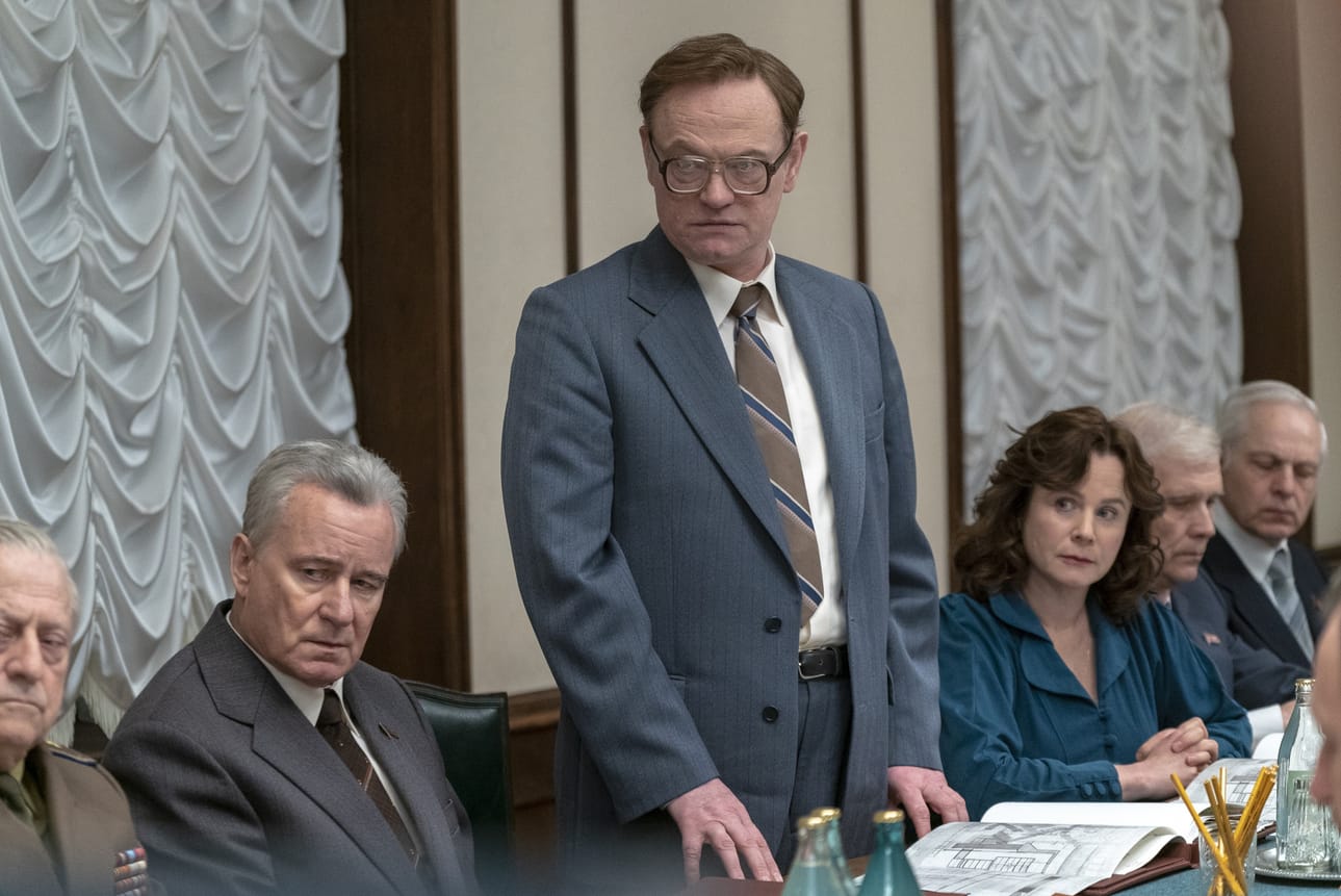 Valeri Legasov (Jared Harris) selvittää seisoaltaan Neuvostoliiton korkeimmalle johdolle, mitä Tshernobylissä on tapahtunut. Chernobyl-sarjan kohtauksessa Boris Shcherbina (Stella Skargård) istuu hänen vasemmalla ja Ulana Homjuk (Emily Watson) oikealla puolellaan.