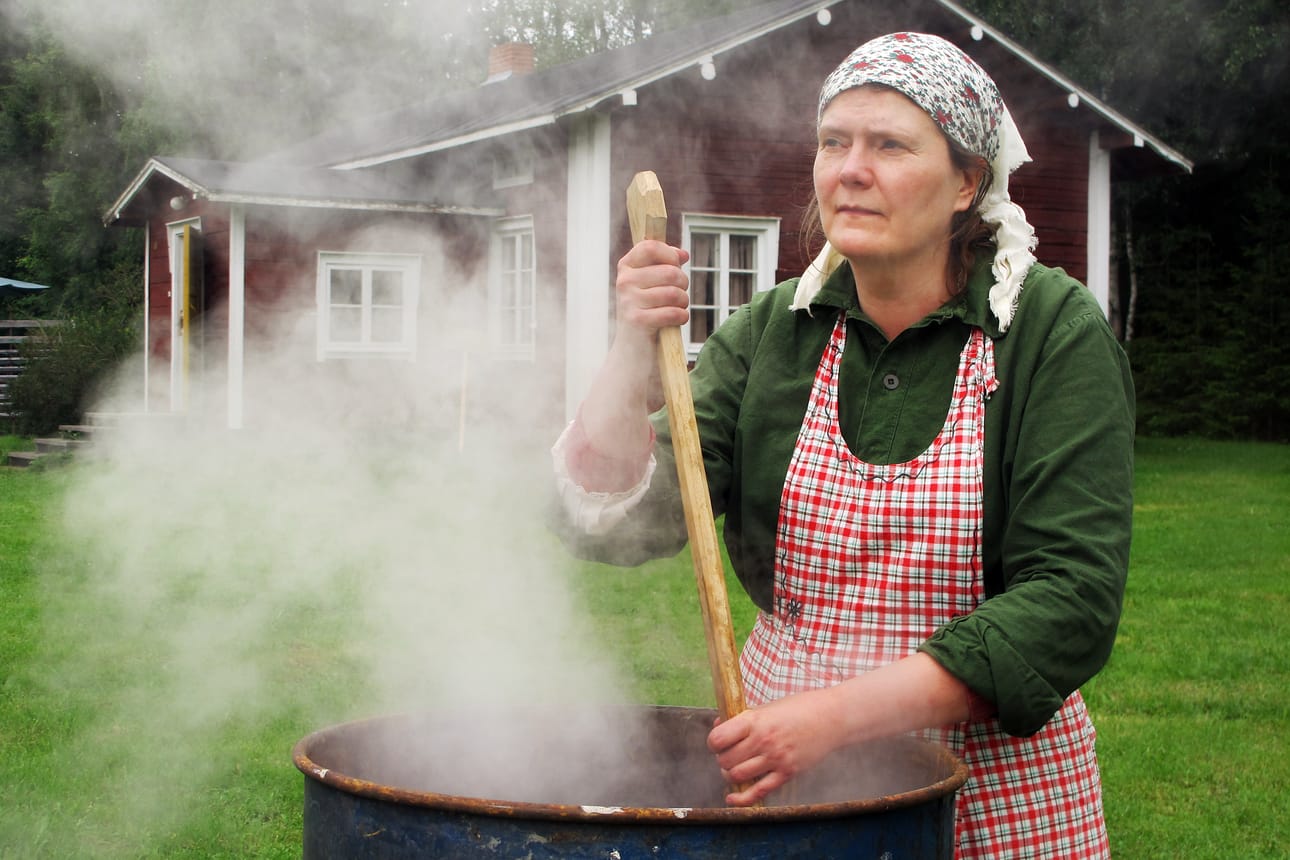 Jaana Vähäsöyrinki keittää maalia aittarakennukseensa. Tällä kertaa värinä on punamulta. Aiemmin Vähäsöyringit ovat keittäneet myös keltamultamaalia. Taustalla näkyy Nivalan kotiseutumuseon Katvalan pieni puoli, jonka seinissä on punamultaa.