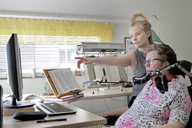 MS-tautia poteva Marketta Haapala selailee kirjoittamiaan runoja yhdessä avustajansa Hanna Rinta-Tassin kanssa. Tietokoneen vierellä sijaitsevat kirjateline ja mekaaninen lukukone, joilla Haapala pystyy selaamaan kirjoja ja lehtiä itsenäisesti. KUVA: Sakari Hällfors