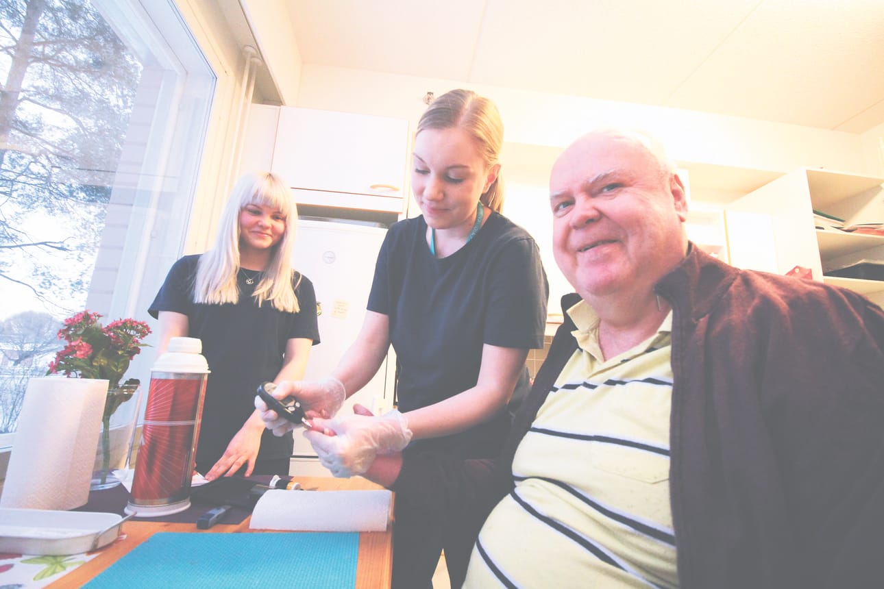 Lähihoitaja Sara Kekkonen (vasemmalla) seuraa, kun työkaveri Mari-Emilia Bräysy mittaa Veikko Toivaisen verensokeria.