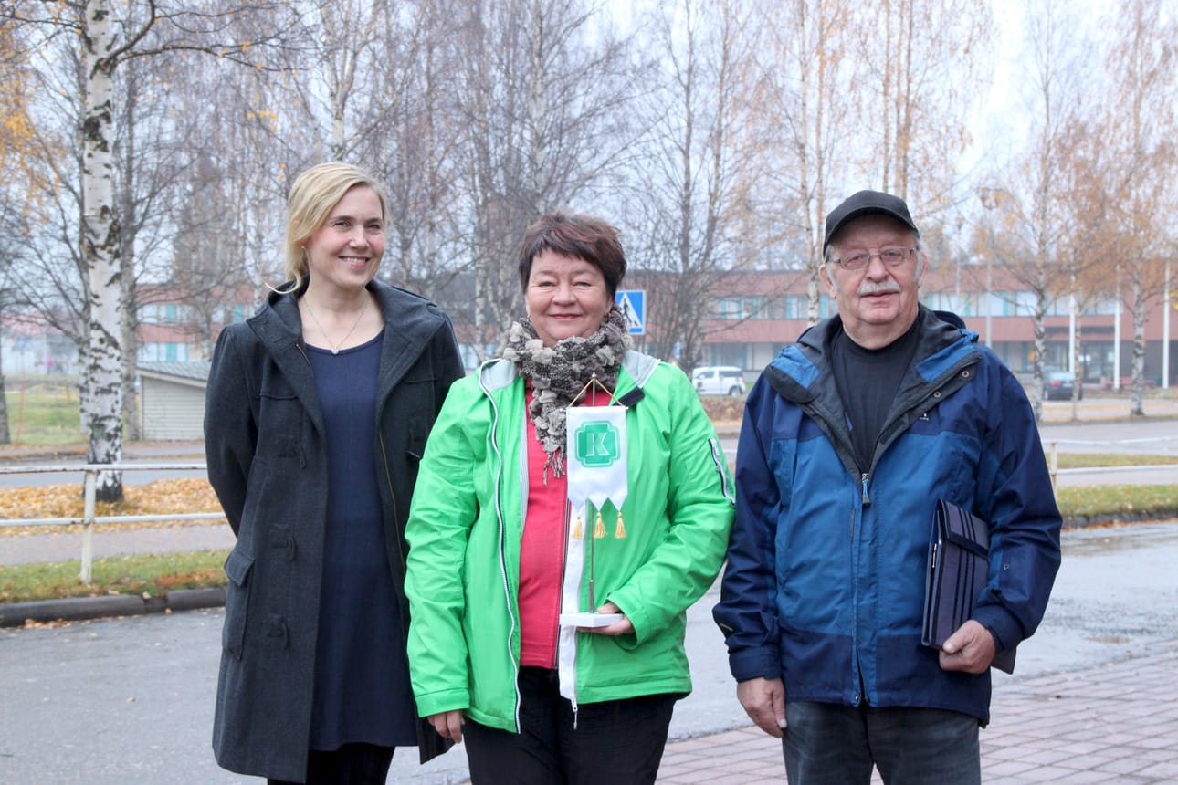 Juhlatoimikunnan jäsenet Mari Kälkäjä, Mirja Luusua ja Martti Räisänen heiluttavat keskustan vihreää apilaviiriä. Juhlatunnelma alkaa nousta.