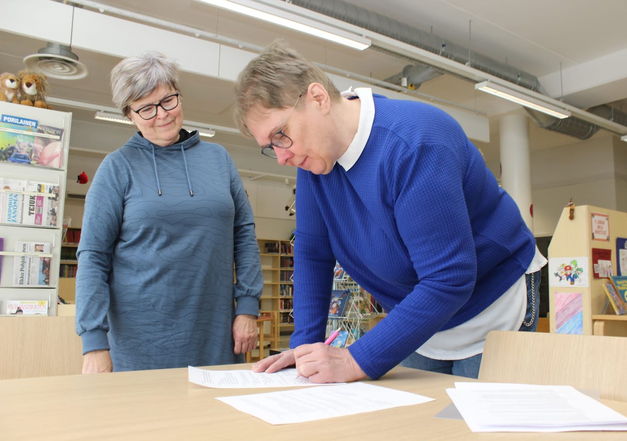 Maj-Britt Uusitalo (taustalla) seuraa, kun kirjastonhoitaja Anna-Maija Haaranoja allekirjoitti paperit Karijoen kunnan puolesta. Sopimus solmittiin tänä aamuna.