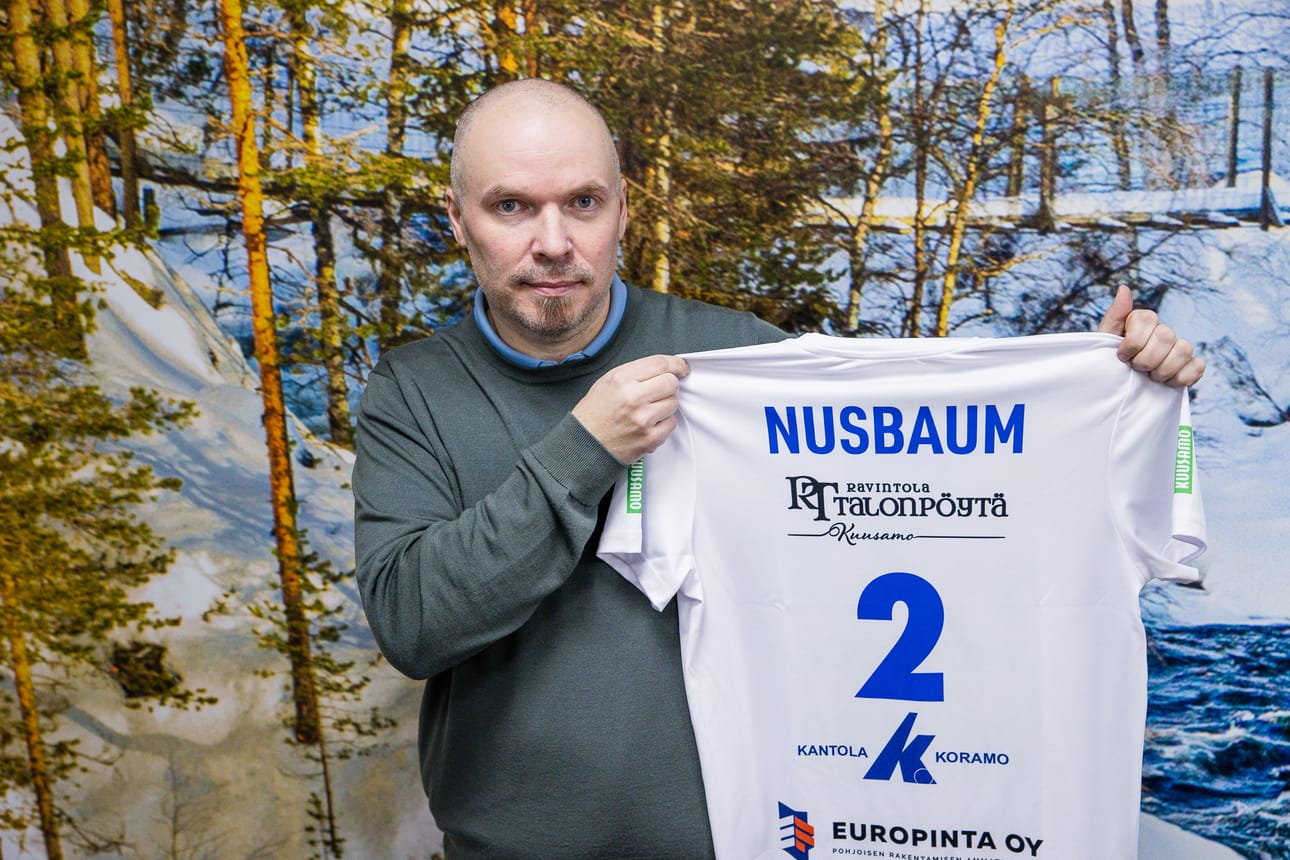Pölkky Kuusamon pelaajakoordinaattori Mika Määttä esittelee joukkueeseen palaavan Carlyle Nusbaumin uunituoretta pelipaitaa.