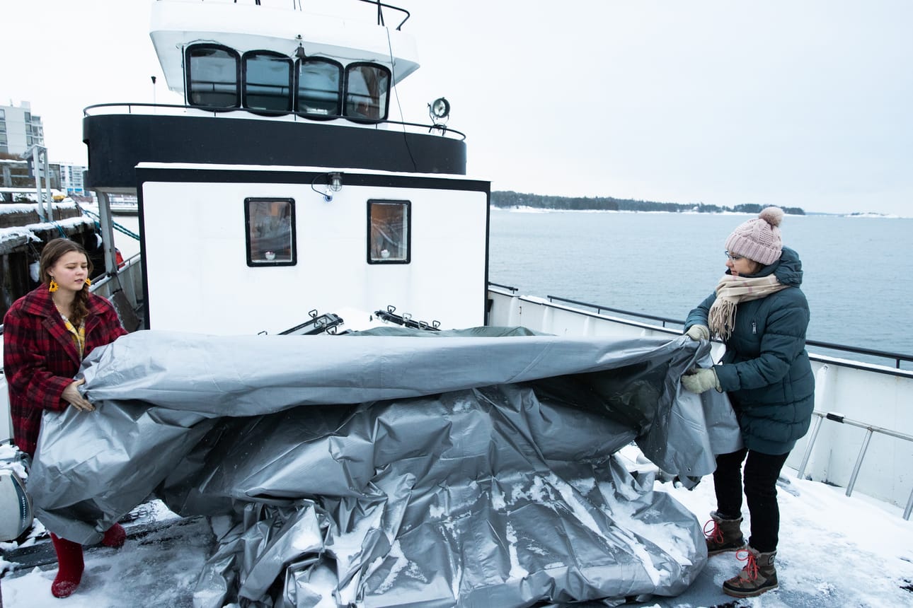 Kova merenkäynti riepottelee toisinaan Helsingin Aurinkolahden satamassa komeilevaa asuntolaiva m/s Suukkoa. Meilläkin, kokeneilla laivureilla, on välillä vaikeaa pysyä pystyssä. Sijainnin vuoksi tänne ei aina pysty ottamaan asiakkaita, kertoo laivassa työskentelevä Mari Korhonen. Julia Korhonen on kaverina siirtämässä pressua pois ruuman kattoikkunoiden yltä.