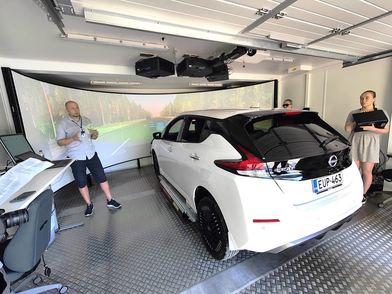 Nissan Leaf on ajettu testipenkkiin ja kuljettajan eteen heijastettu aidolta näyttävä maisemakuva. Testiolosuhteet vastaavat mahdollisimman tarkasti aitoja liikennetilanteita, apulaisprofessori Tuomo Kujala ja tutkimusavustaja Tuulia Verlin esittelevät. Kuva: Kari Pitkänen