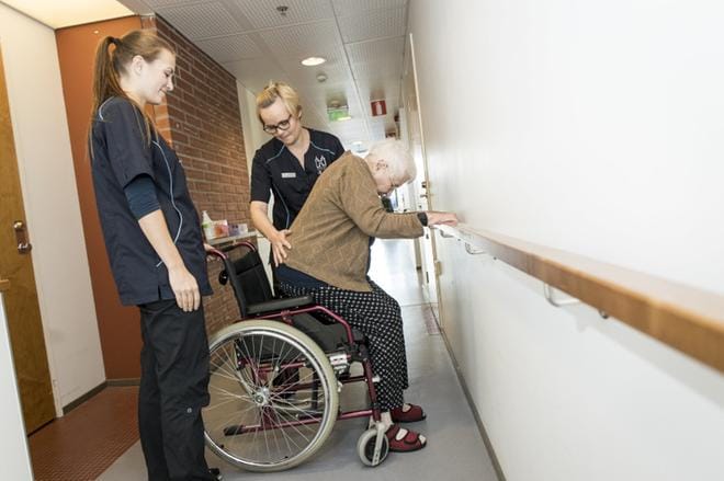 Fysioterapeutti Suvi Saukkola ja lähihoitaja Riikka Koivuranta-Hakala auttavat Riitta Siikasta nousemaan pyörätuolista hyvinvointikeskus Keinukamarin käytävällä. Kyseessä ei ole testitilanne. KUVA: PEKKA RAUTIAINEN