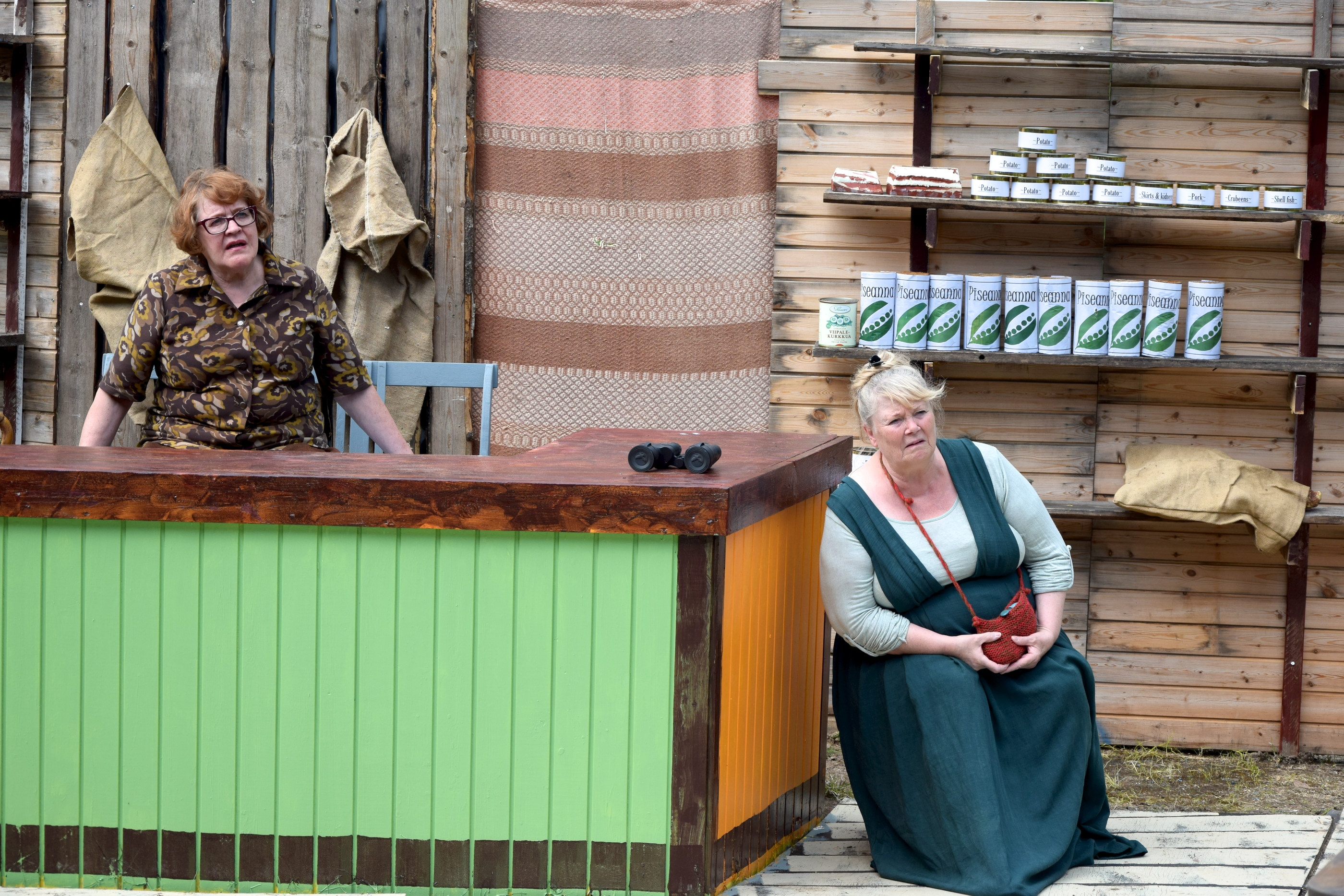 Eileen (Leena Lanne, vasemmalla) ja Kate (Elvi Tapia Olguin) kuuntelevat kiinnostuneina Johnnypatmiken (Jyrki Hyvölä) uutisten lukua kaupassaan.