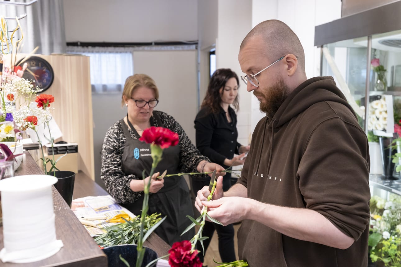 Toimittaja Henri Ruuska pääsi trimmaamaan neilikoita surulaitteita varten floristimestari Minna Heikkilä-Tuovilan (takana vas.) kanssa. Taustalla myös Maria Kajava.