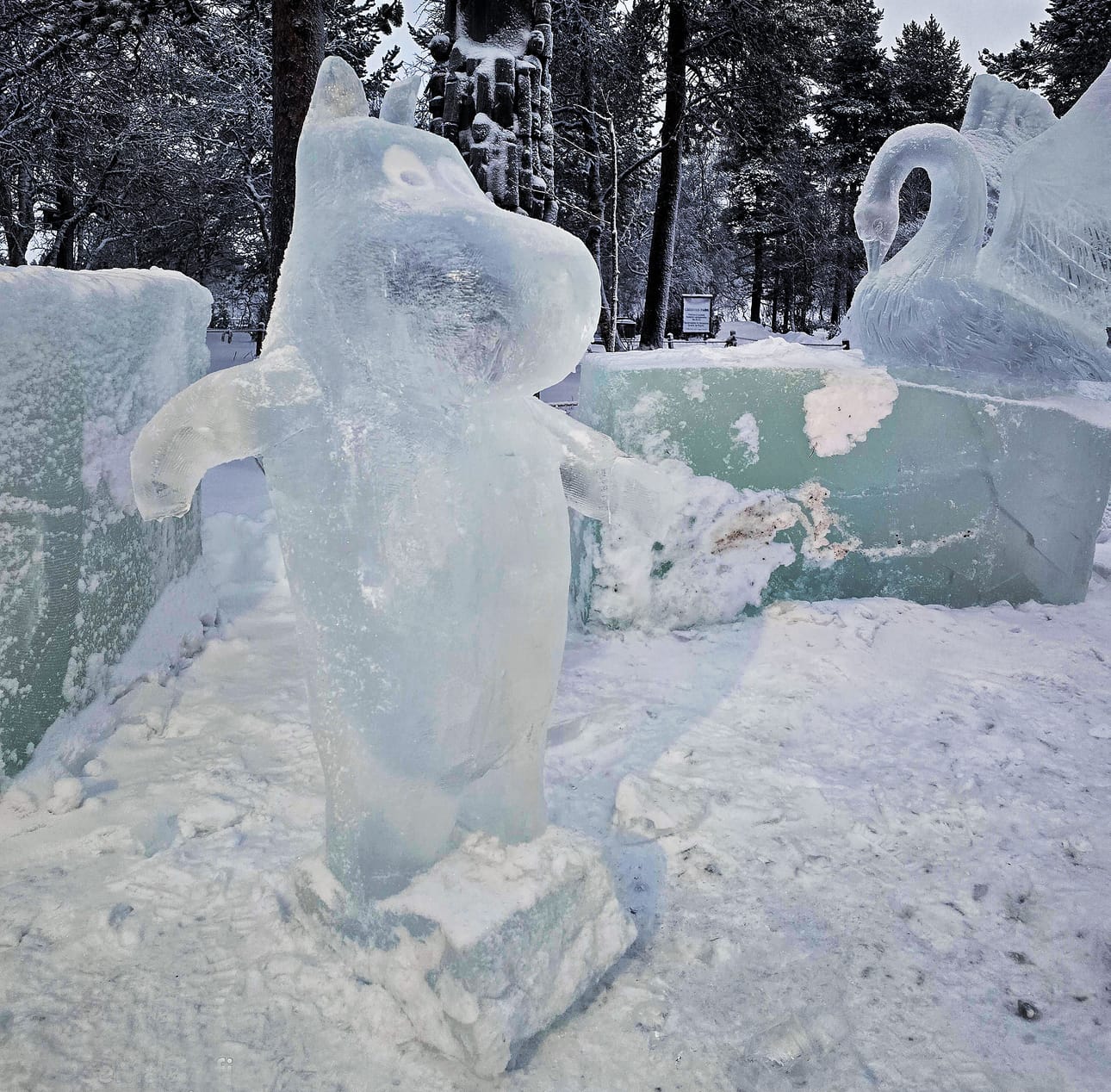 Jäästä veistetty Muumipeikko tervehtii ohikulkijoita Kiilopäällä. Taustalla talvehtii jäinen joutsen.