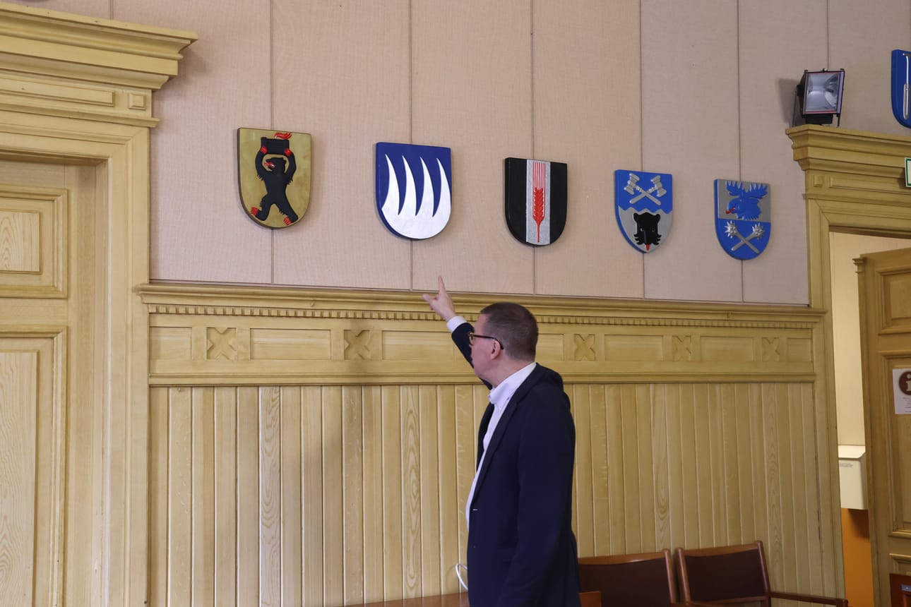 Kaupunginjohtaja Markku Lumio Kaskisten komeassa valtuustosalissa, jonka seinää koristavat puiset kuntavaakunat. Lumio viittoilemassa Karijoen vaakunan suuntaan. Karijoki on yksi neljästä paikkakunnasta, jonka kuntaliitoshalukkuutta Lumio haluaa tiedustella.