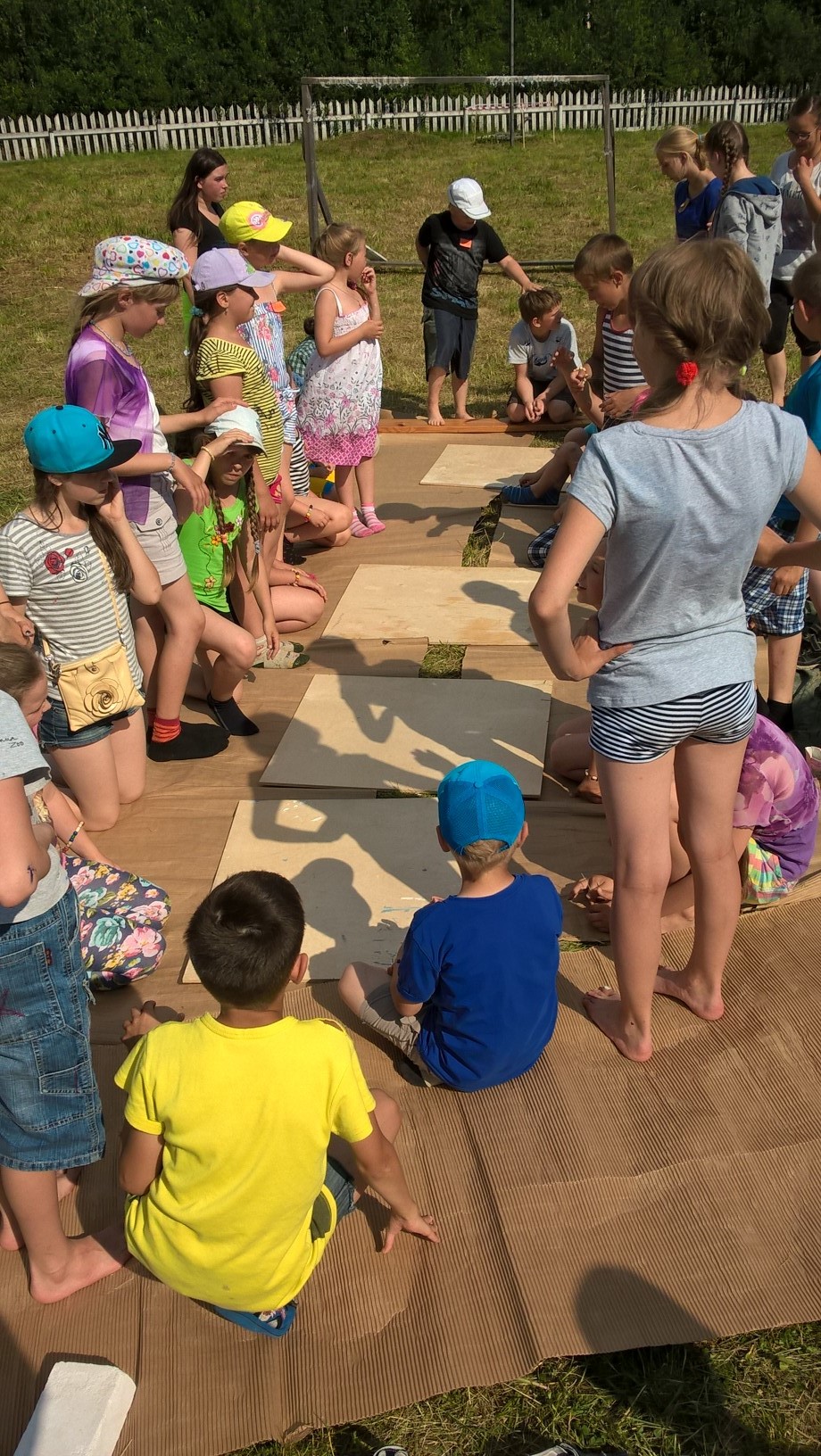 Leiriläiset valmistumassa pihanurmikolla kahteen sisäkkäiseen piiriin, joissa ulompana istuvat piirsivät sisempänä istuvan naamakuvan paperilautasen takapuolelle. Kun yksi osa oli piirretty, piirtäjä siirtyi seuraan eteen lisäämään jonkun muun kasvon osan.